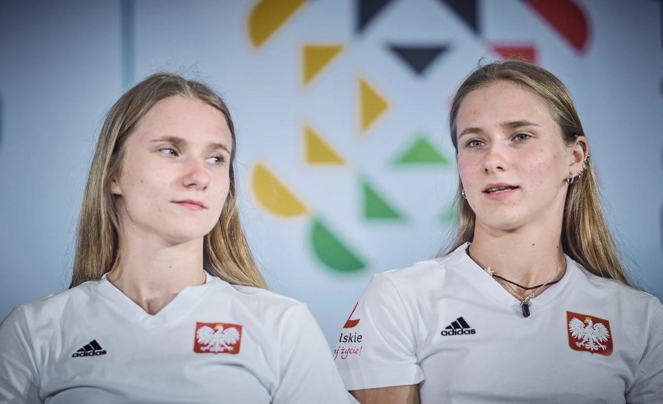 Aleksandra i Natalia Kałucka Dwie młode kobiety ubranej w białe koszulki sportowe z godłem Polski oraz logotypami sponsorów, siedzą obok siebie na tle rozmytego, kolorowego wzoru. Jedna z nich patrzy w kierunku drugiej.