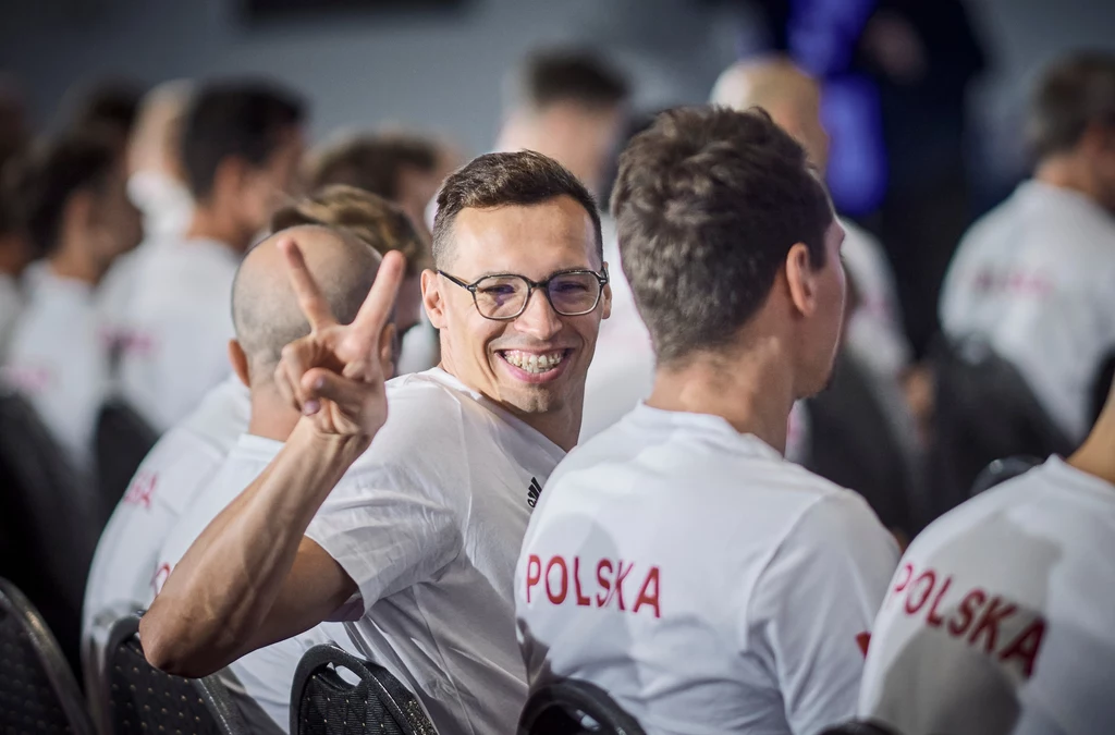 Mężczyzna w okularach z szerokim uśmiechem siedzi w grupie osób ubranych w białe koszulki z napisem POLSKA na plecach, pokazując znak pokoju palcami przed obiektywem.