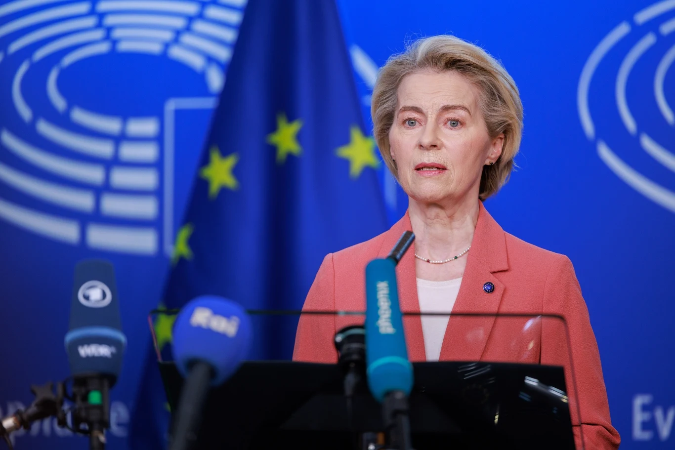 Głosowanie nad wotum nieufności dla KE, którą kieruje Ursula von der Leyen odbędzie się w czwartek w Strasburgu Kobieta w jasnej marynarce stoi przed mikrofonami podczas konferencji prasowej, w tle widoczne symbole Unii Europejskiej oraz charakterystyczna niebieska flaga z gwiazdami.