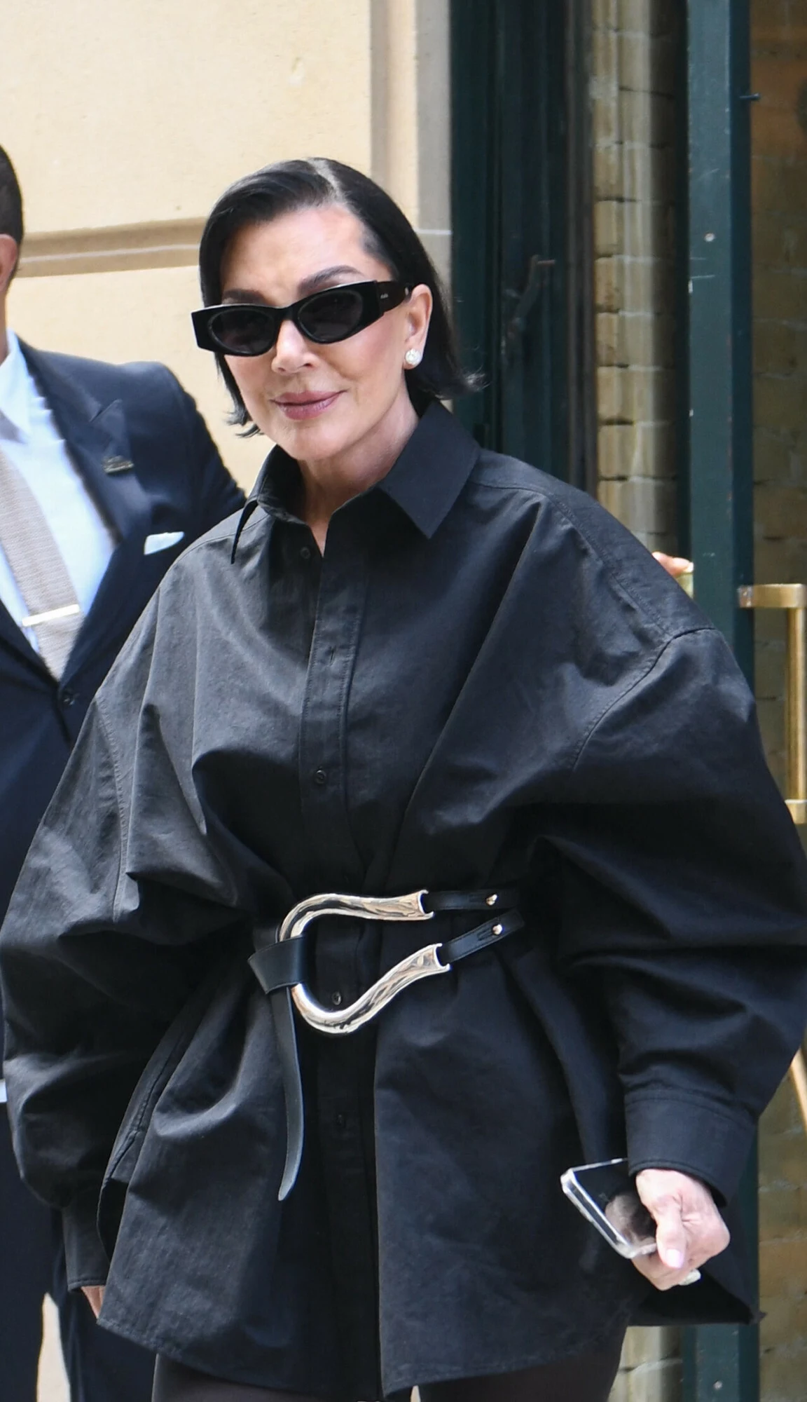 Kris Jenner zaprezentowała twarz po liftingu Elegancka kobieta w czarnych okularach przeciwsłonecznych oraz oversizowej, czarnej koszuli z szerokim paskiem o srebrnej klamrze, trzymająca telefon, stoi w pobliżu wejścia do budynku.