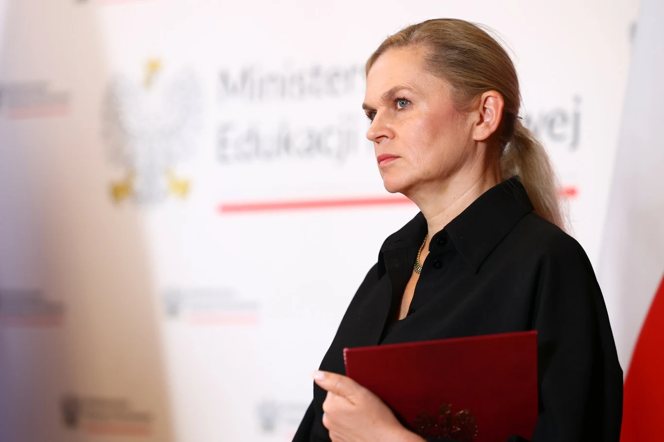 Barbara Nowacka ujawniła datę nowej matury i egzaminu ósmoklasisty Kobieta w ciemnym stroju trzymająca w rękach czerwony dokument, w tle logo Ministerstwa Edukacji oraz fragment flagi narodowej.