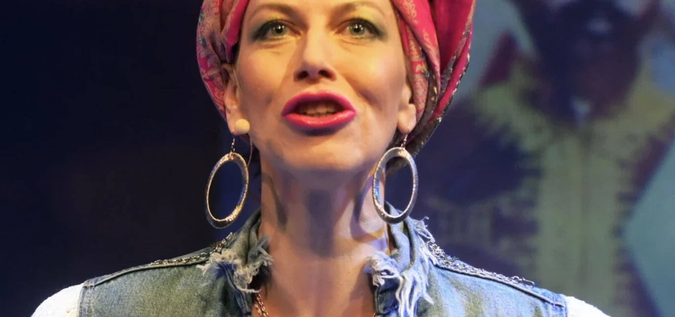 Katarzyna Chlebny jako Rita Marley Kobieta z dużymi kolczykami, w różowej chuście na głowie oraz dżinsowej kamizelce, patrzy do przodu w teatralnym oświetleniu.