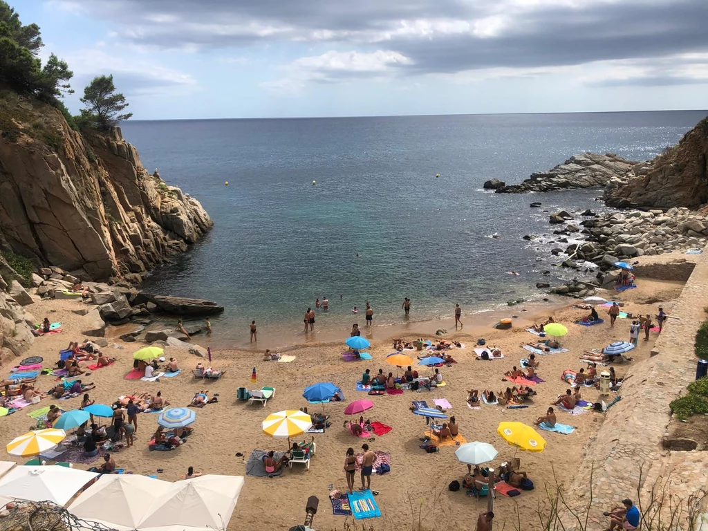 Barcelona Piaszczysta plaża otoczona skalistymi klifami, na której wypoczywa wiele osób pod kolorowymi parasolami; część ludzi kąpie się w spokojnej, błękitnej wodzie zatoki, a w tle rozciąga się spokojne morze pod częściowo pochmurnym niebem.