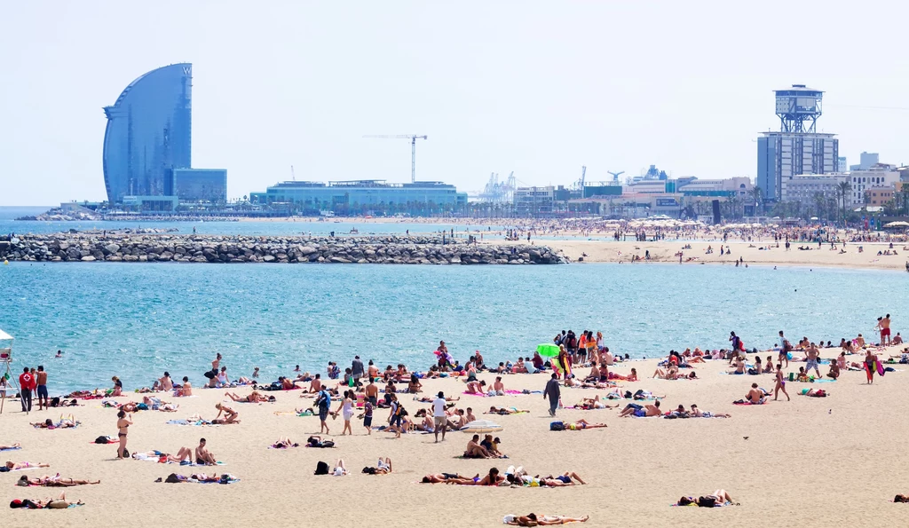 Barcelona Plaża pełna ludzi wypoczywających na piasku, w tle spokojne morze, nowoczesne budynki i charakterystyczny hotel w kształcie żagla.