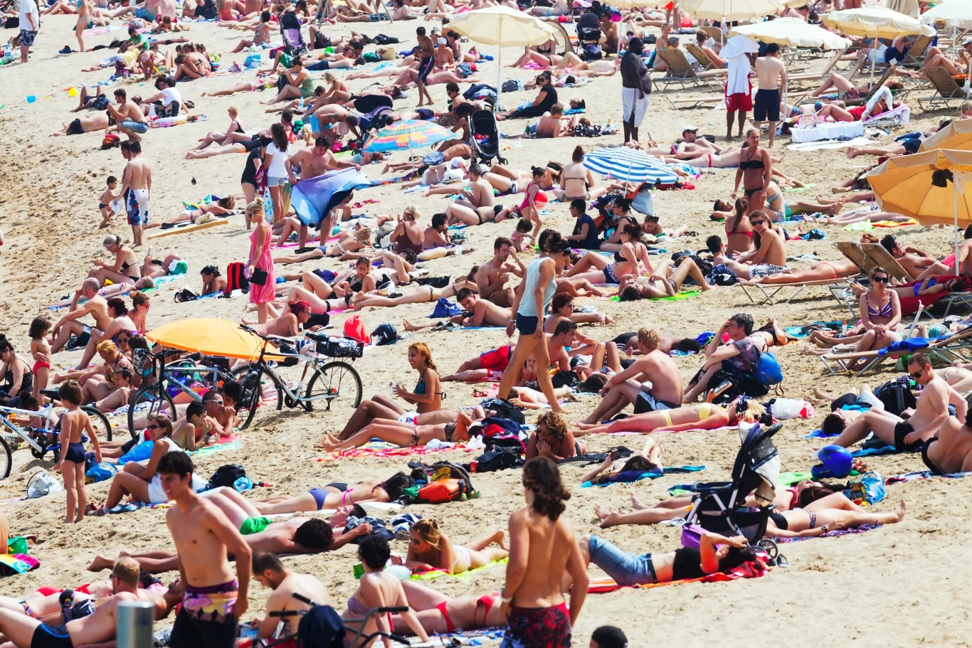 Tłum ludzi opalających się i odpoczywających na zatłoczonej plaży, część osób leży na ręcznikach lub leżakach, a niektórzy spacerują po piasku, w tle widoczne kolorowe parasole przeciwsłoneczne i rowery.