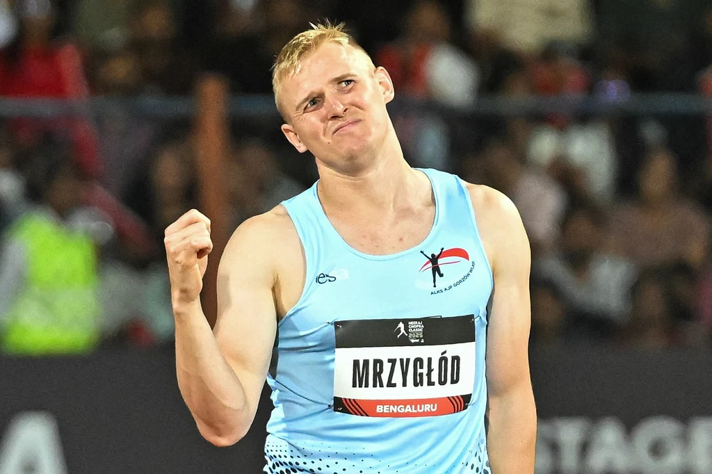 Cyprian Mrzygłód Sportowiec w błękitnym stroju lekkoatletycznym z zaciśniętą pięścią i wyrazem zdecydowania na twarzy, z tabliczką z nazwiskiem na piersi, na tle rozmytego tłumu kibiców na stadionie.