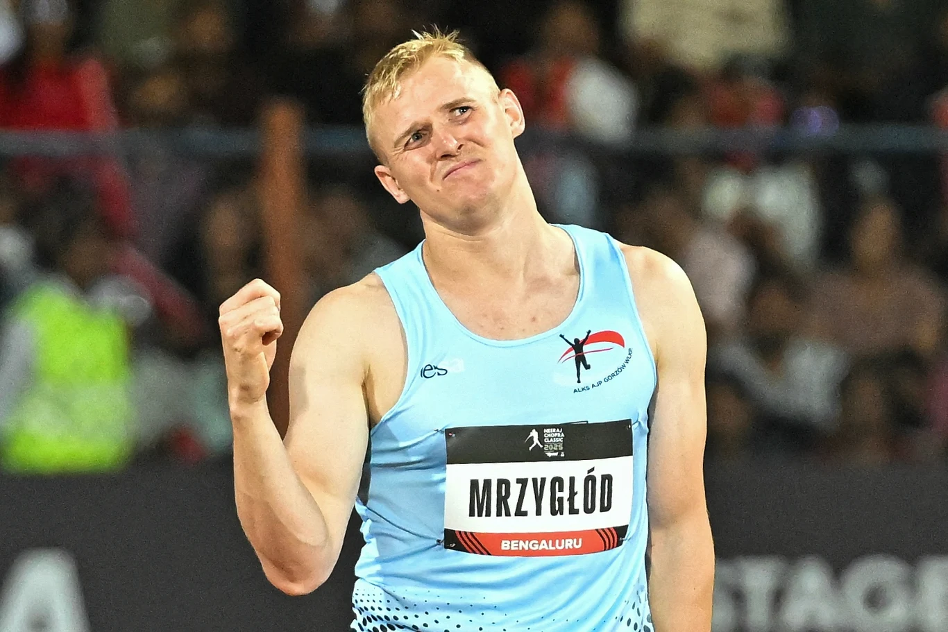 Cyprian Mrzygłód Sportowiec w błękitnym stroju lekkoatletycznym z zaciśniętą pięścią i wyrazem zdecydowania na twarzy, z tabliczką z nazwiskiem na piersi, na tle rozmytego tłumu kibiców na stadionie.