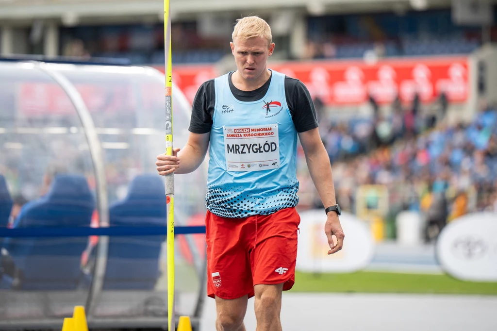 Zawodnik lekkoatletyczny przygotowujący się do rzutu oszczepem na stadionie, ubrany w sportowy strój z numerem startowym i nazwiskiem, w tle widownia oraz fragment trybun.