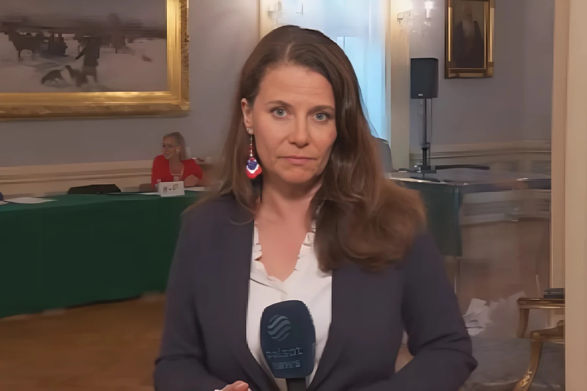 Dziennikarka w granatowej marynarce z mikrofonem w dłoni stoi w eleganckim wnętrzu z zielonym stołem konferencyjnym w tle, na ścianach wiszą obrazy, a przy stole siedzi kobieta.