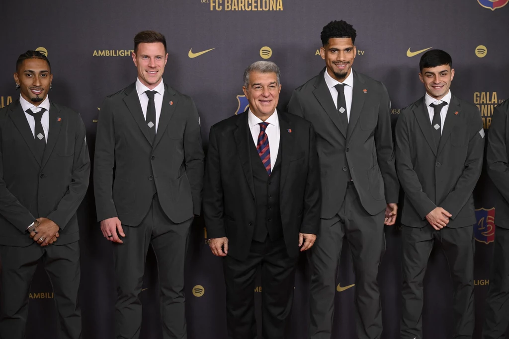 Raphinha, Marc-Andre ter Stegen, Joan Laporta, Ronald Araujo, Pedri Grupa pięciu elegancko ubranych mężczyzn stoi na tle ścianki z logo FC Barcelona, wszyscy uśmiechnięci, ubrani w szare garnitury z krawatami, jeden z mężczyzn pośrodku wyróżnia się czarnym garniturem i kolorowym krawatem.