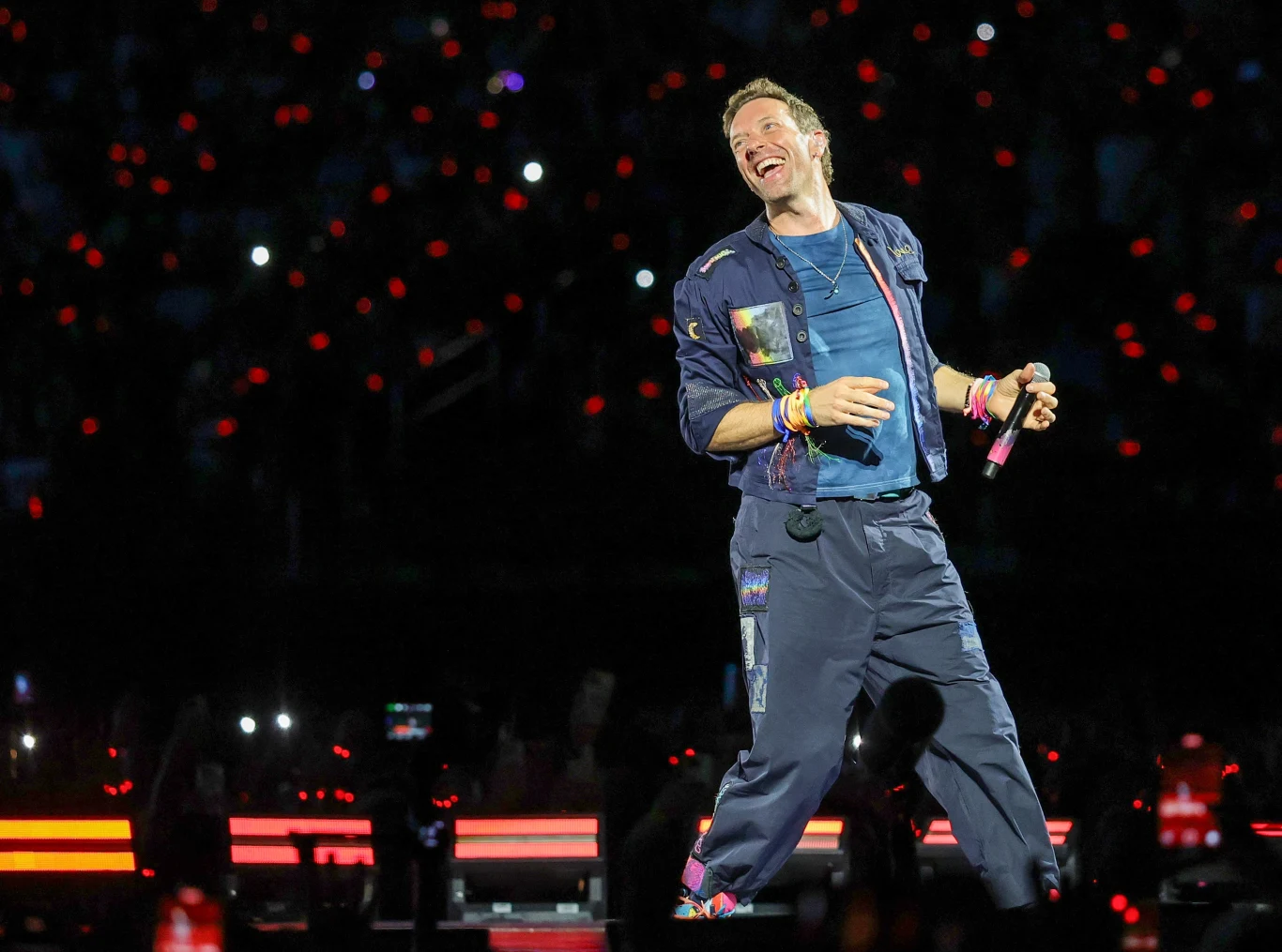 Coldplay świętuje 25-lecie albumu "Parachutes" Wokalista na scenie koncertowej trzyma mikrofon, ubrany w kolorowe ubrania, wokół niego świecą opaski na nadgarstkach i światła publiczności tworzące atmosferę widowiska.