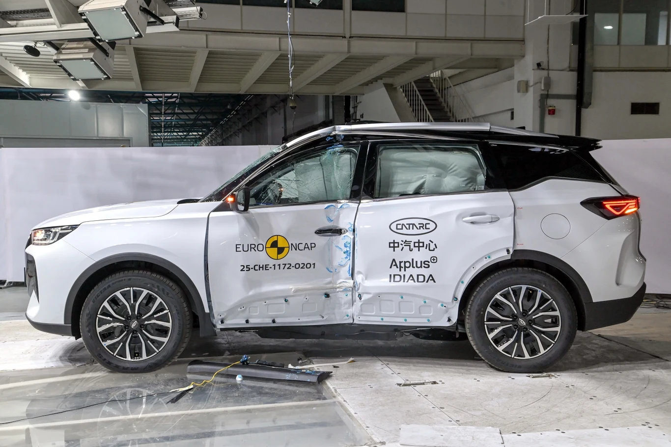 Biały samochód typu SUV z widocznymi uszkodzeniami na drzwiach, logo Euro NCAP i innymi oznaczeniami instytutów testujących, ustawiony w specjalistycznym pomieszczeniu podczas testu zderzeniowego.
