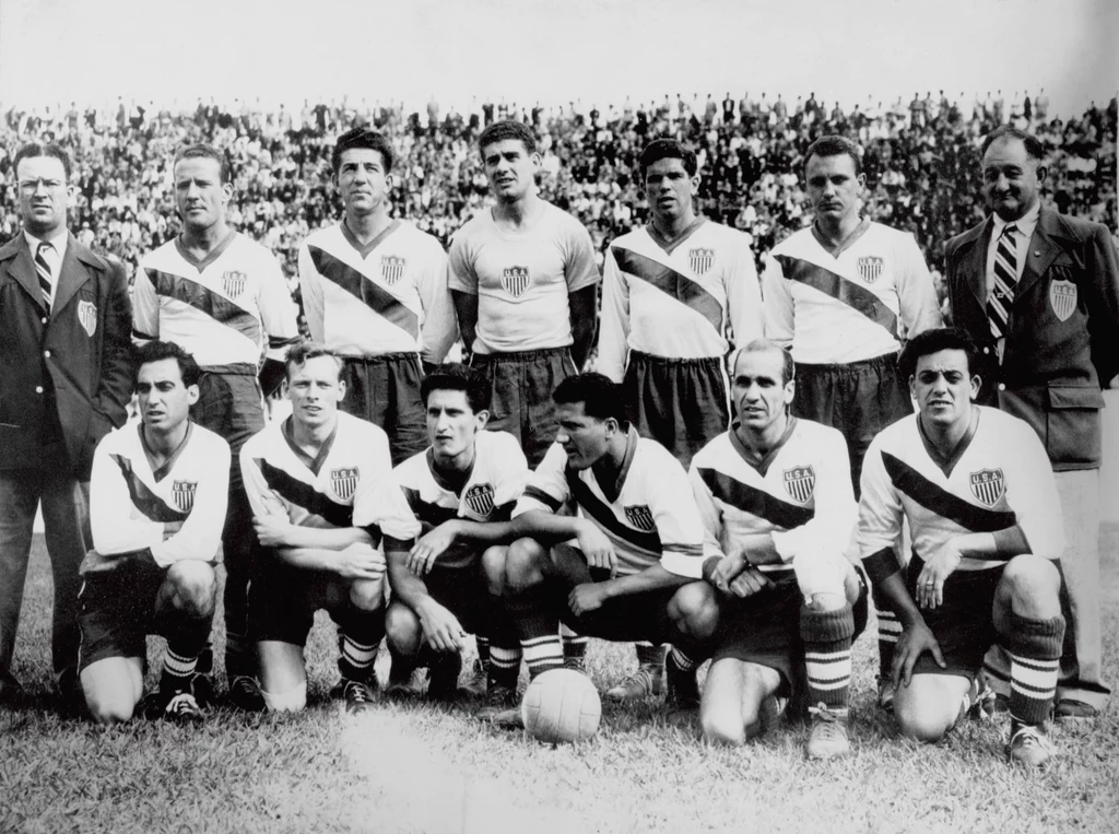 Reprezentacja USA na mundial w 1950 roku. Joe Gaetjens (trzeci od prawej w dolnym rzędzie) Członkowie drużyny piłkarskiej ustawieni w dwóch rzędach na boisku, wszyscy ubrani w jednolite stroje sportowe z charakterystycznym ukośnym pasem oraz herbem na piersi, za piłkarzami widać tłum kibiców na trybunach.