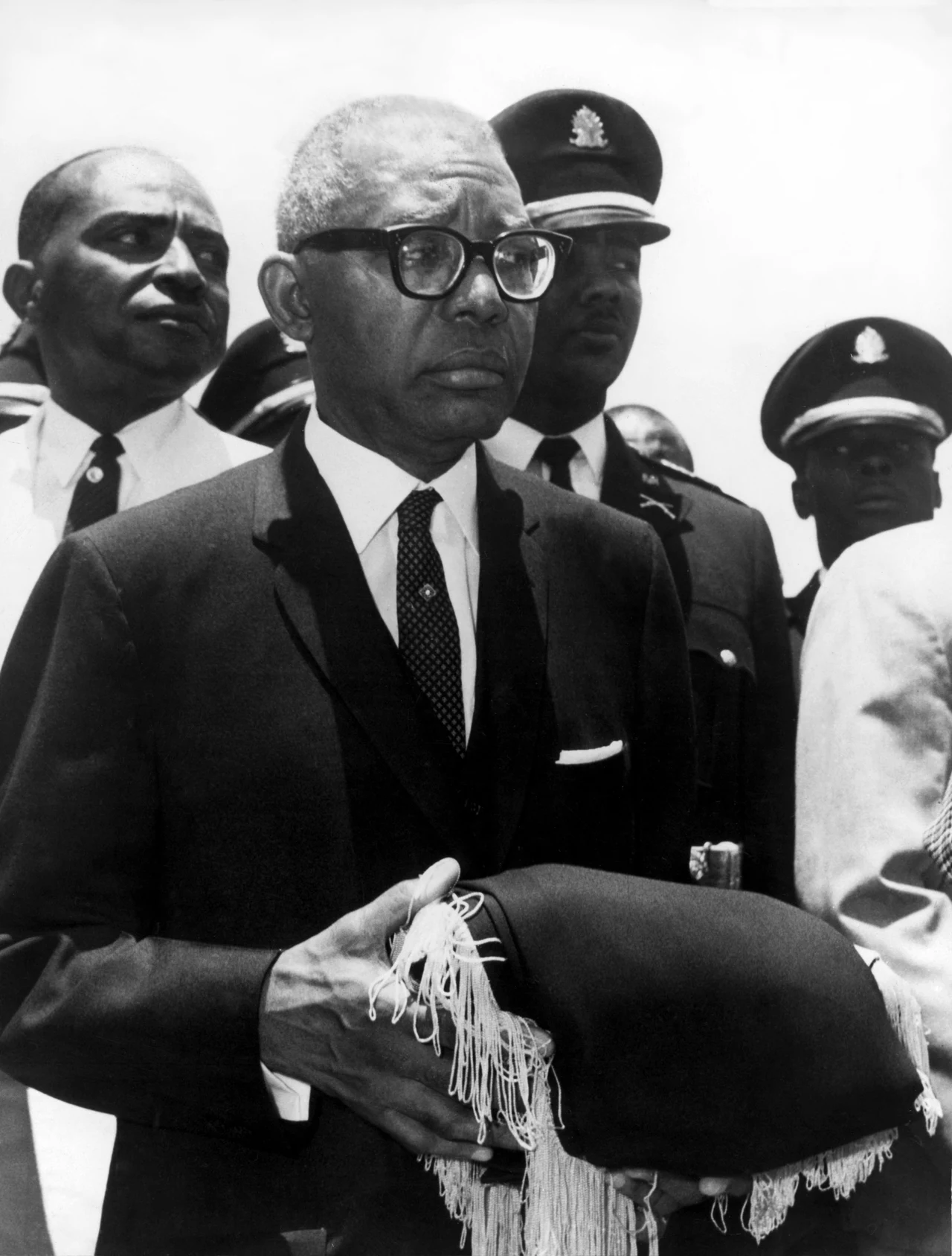 Francois Duvalier Starszy mężczyzna w garniturze i okularach trzyma ozdobioną frędzlami togę, otoczony przez kilku mężczyzn w mundurach policyjnych.