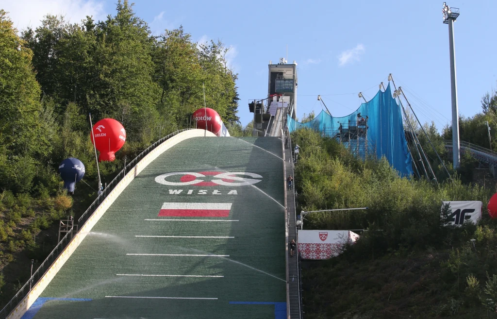 Skocznia w Wiśle-Malince podczas konkursu Letniego Grand Prix Skocznia narciarska otoczona zielonymi drzewami oraz balonami reklamowymi, widoczne są oznaczenia na rozbiegu, polska flaga oraz napis Wisła, w tle budynek sędziowski.