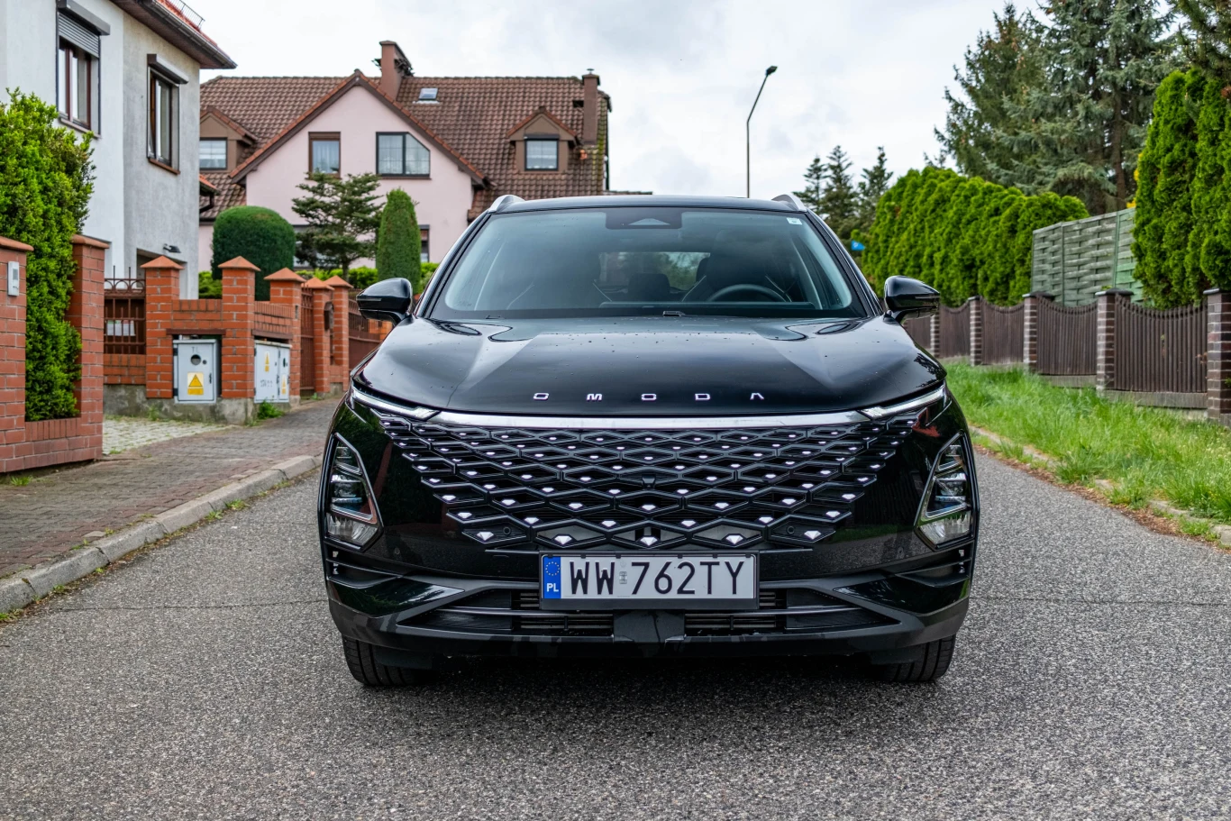 Z danych IBRM SAMAR wynika, że dla wielu polskich nabywców chiński samochód może być pierwszym nowym autem w życiu Czarny samochód osobowy marki OMODA stoi zaparkowany przodem do widza na spokojnej ulicy osiedlowej, po bokach widoczne są domy jednorodzinne, ogrodzenia i zielone drzewa.