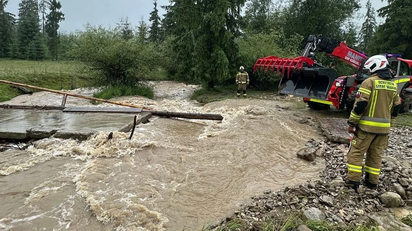 Potok zablokował wodę w rejonie Witowa Strażacy w odblaskowych mundurach przy uszkodzonym moście nad wzburzonym potokiem, wokół rozlana woda i pojazd strażacki z wysięgnikiem, całość w otoczeniu zieleni i drzew.