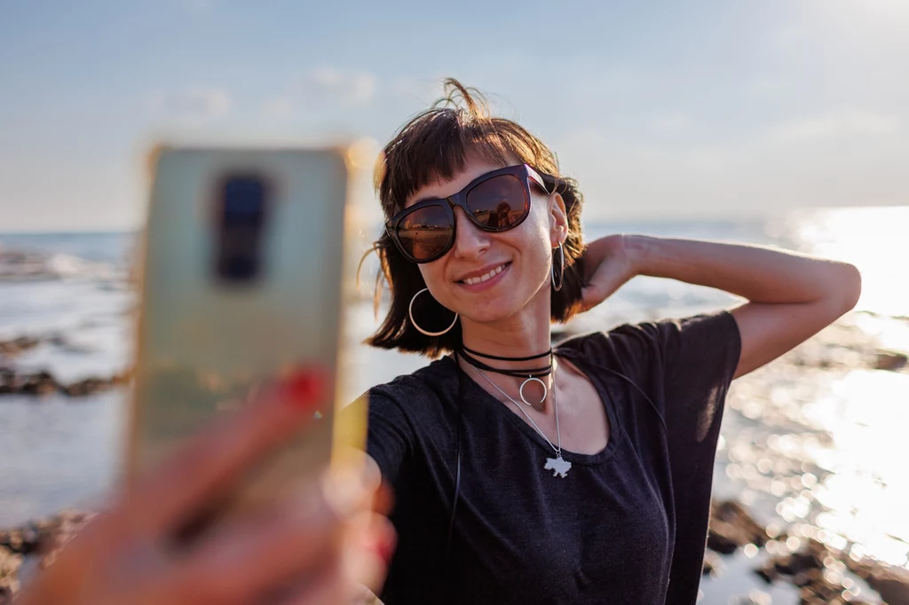 15 proc. goli pachy okazjonalnie – na lato lub przed ważnym wydarzeniem Kobieta w ciemnych okularach przeciwsłonecznych robi sobie selfie nad brzegiem morza, w tle widoczne są fale oraz jasne niebo.