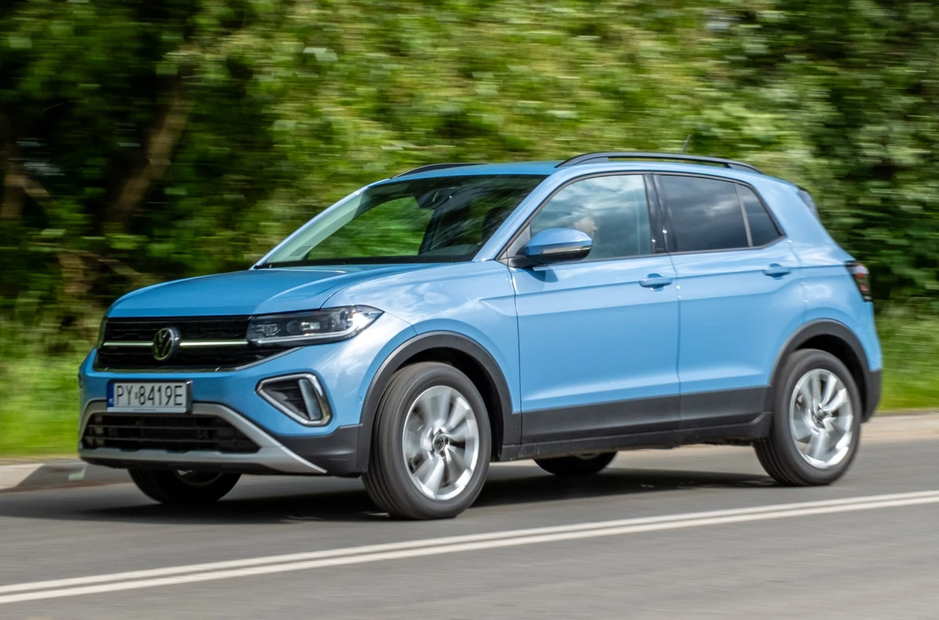 Wyposażenia T-Crossa, auta bądź co bądź głównie miejskiego, jeszcze do niedawna można było spotkać głównie w większych i droższych modelach. W T-Crossie większość oczywiście wymaga dopłaty. Niebieski samochód osobowy marki Volkswagen SUV jedzie po asfaltowej drodze, w tle zielone drzewa i rozmyte otoczenie wskazujące na ruch pojazdu.
