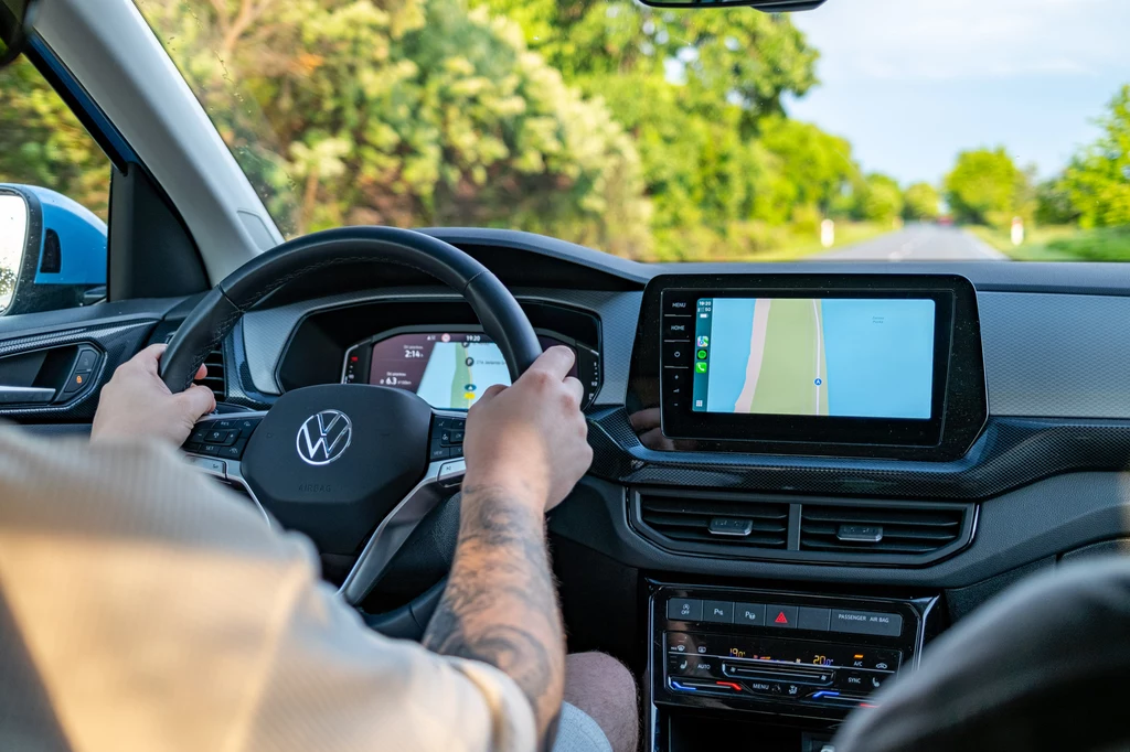 Wnętrze samochodu marki Volkswagen podczas jazdy, kierowca trzyma ręce na kierownicy, na centralnym ekranie wyświetlana jest mapa nawigacji, przez szyby widoczna zielona, letnia sceneria i asfaltowa droga.
