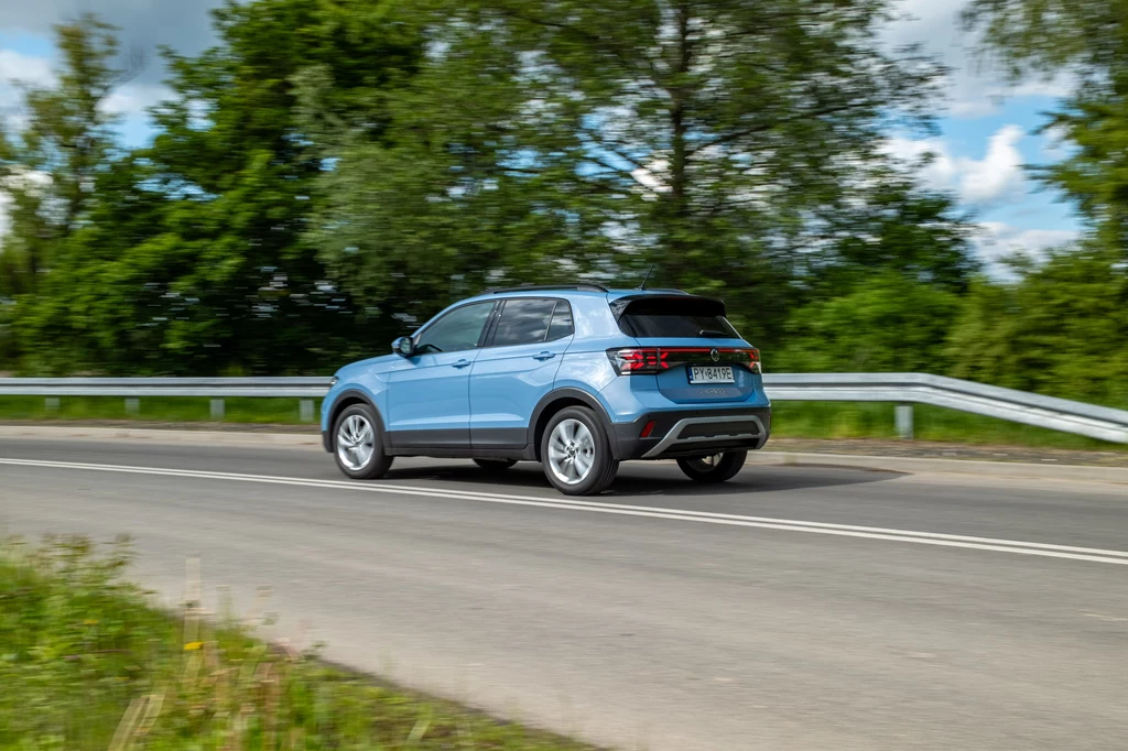 niebieski samochód osobowy typu SUV poruszający się po asfaltowej drodze otoczonej zielenią i barierkami, w tle drzewa oraz pochmurne niebo