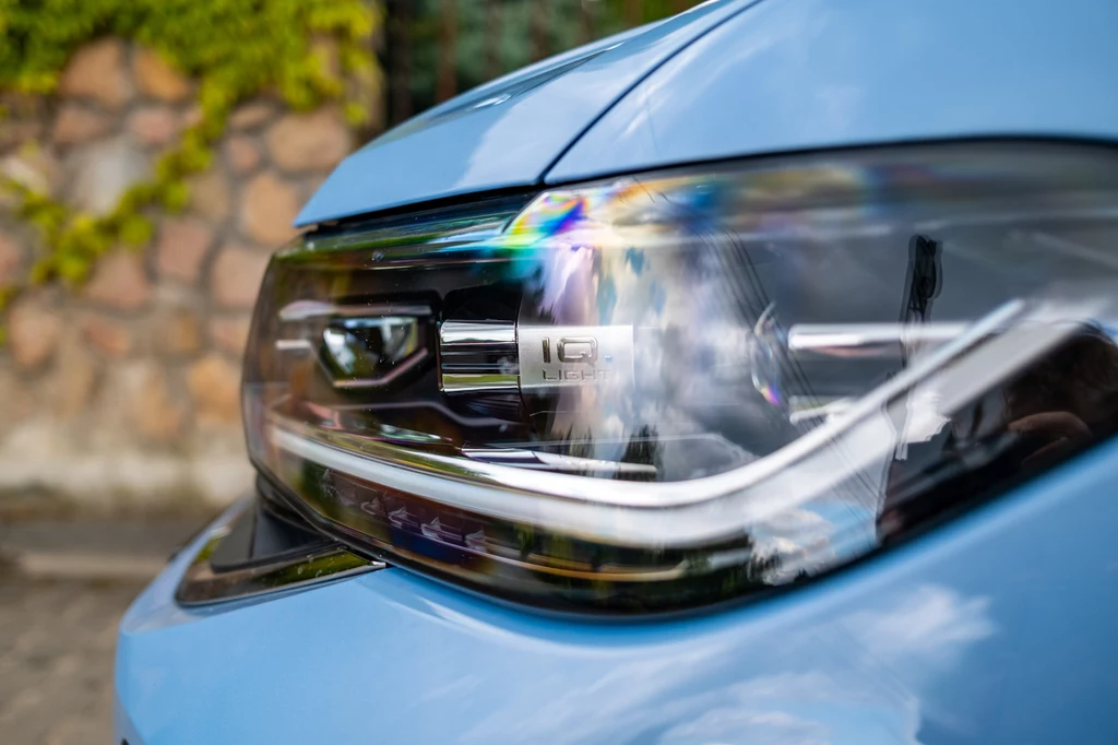 Zbliżenie na przedni reflektor niebieskiego samochodu, z wyraźnymi detalami szklanej obudowy oraz elementów lampy, w tle widoczny fragment murka z kamienia i roślinności.