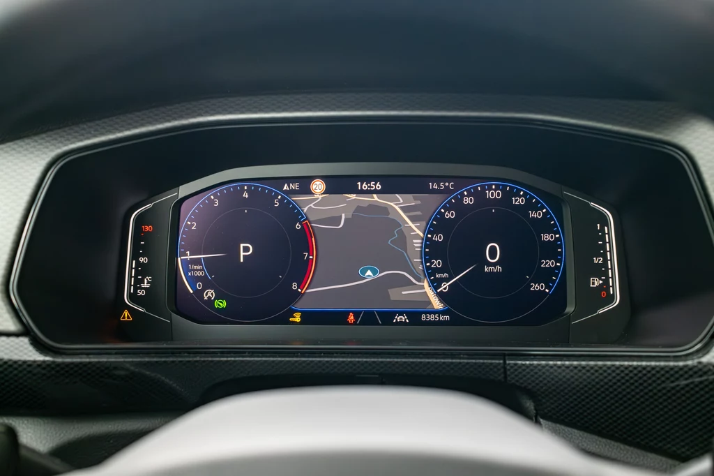 Cyfrowy panel zegarów samochodowych z wyświetlanym na środku ekranem nawigacji GPS, wskaźnikiem prędkości po prawej stronie oraz wskaźnikiem trybu jazdy po lewej. Otaczające elementy mają nowoczesny, czytelny design.