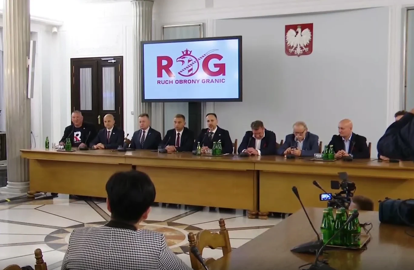 Posiedzenie Parlamentarnego Zespołu Ruch Obrony Granic Grupa mężczyzn w garniturach siedzących przy długim stole konferencyjnym, w tle ekran z napisem Ruch Obrony Granic oraz godło Polski, obecność mikrofonów i kamer sugeruje oficjalną konferencję prasową lub spotkanie polityczne.