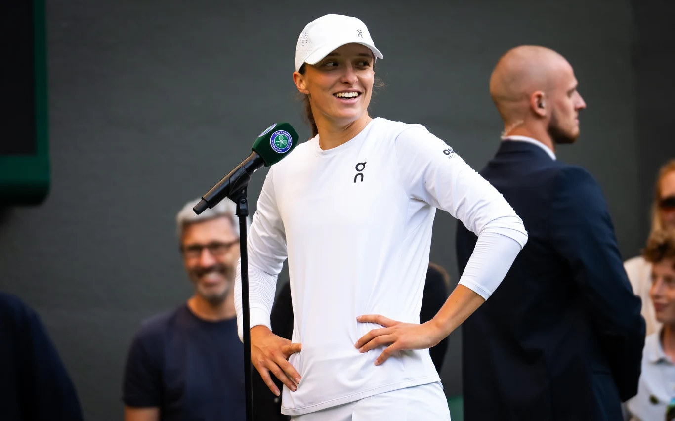 Iga Świątek Tenisistka ubrana w biały strój sportowy oraz czapkę z daszkiem stoi przy mikrofonie na tle publiczności i osób towarzyszących, uśmiechając się podczas wywiadu po meczu.