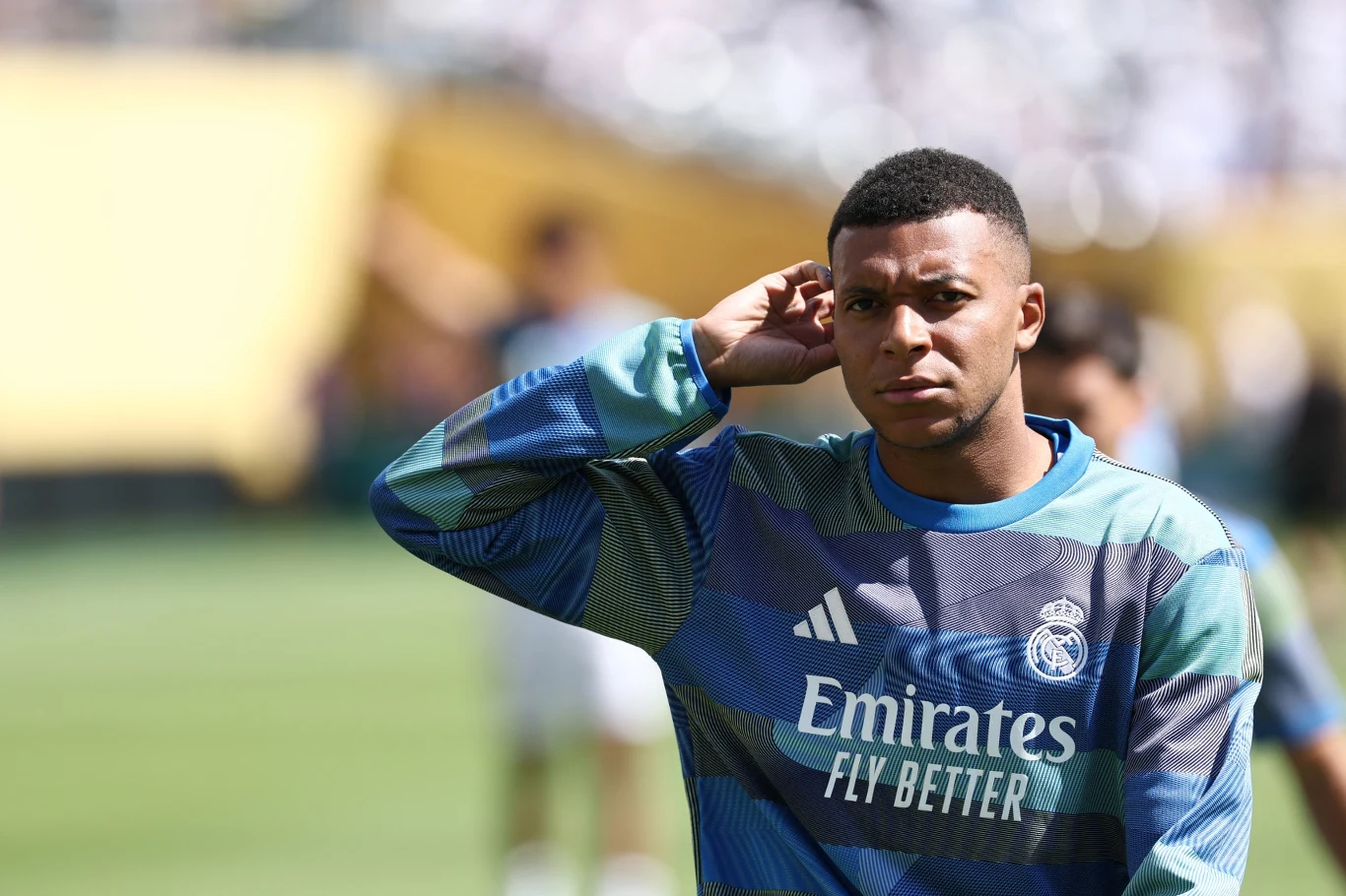 Piłkarz w dresie treningowym Realu Madryt z logo Emirates na stadionie podczas przygotowań lub rozgrzewki, w tle widoczna rozmyta publiczność i trybuny.