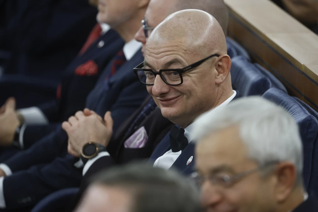 Mężczyzna w garniturze i okularach siedzi wśród innych elegancko ubranych osób na sali, lekko się uśmiecha.