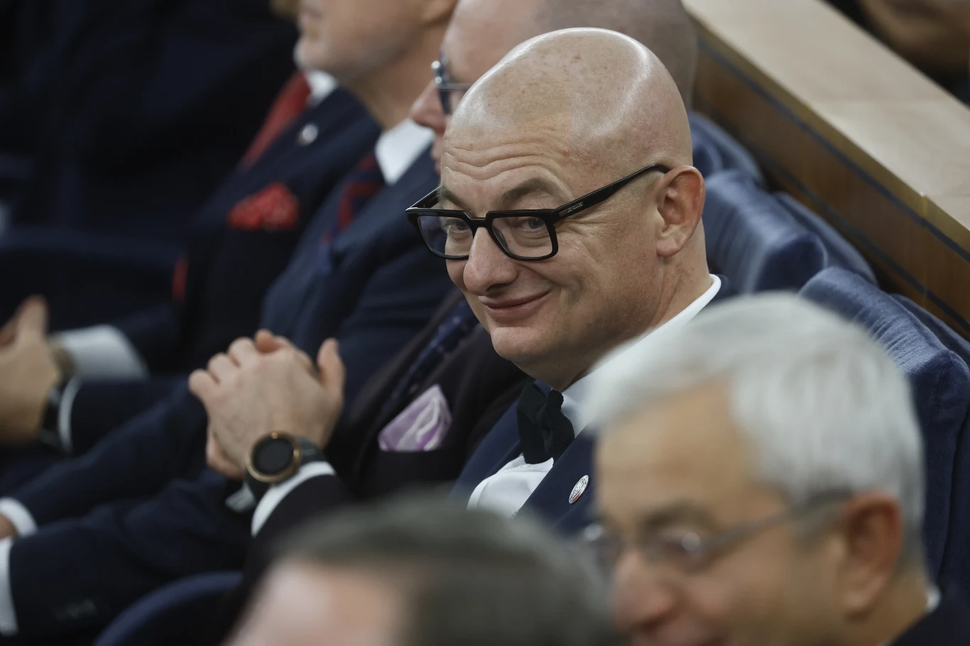 Mężczyzna w garniturze i okularach siedzi wśród innych elegancko ubranych osób na sali, lekko się uśmiecha.