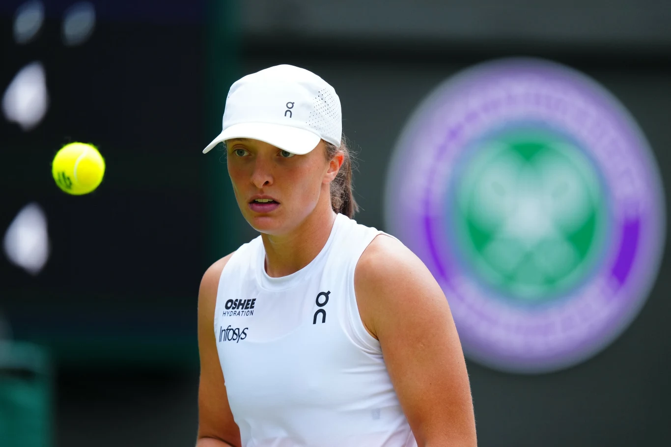 Tenisistka w białym stroju sportowym i czapce z daszkiem koncentruje się na żółtej piłce tenisowej, w tle rozmyte logo turnieju Wimbledon.