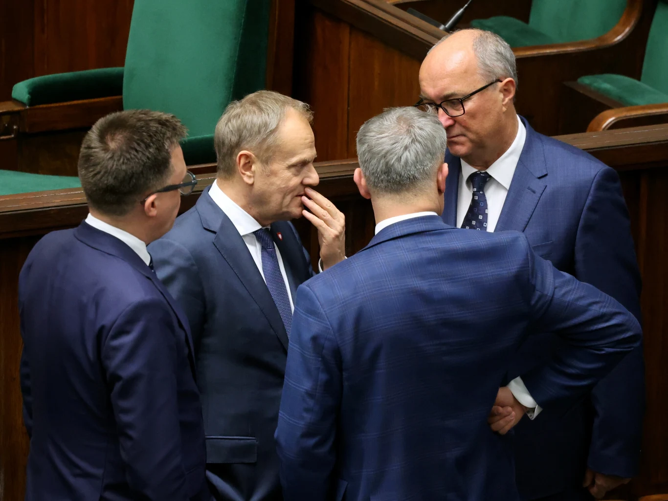 Media ujawniają kulisy spotkania liderów koalicji rządzącej (zdjęcie ilustracyjne) Czterech mężczyzn ubranych w garnitury prowadzi poważną rozmowę stojąc w kręgu w dużej sali z zielonymi fotelami