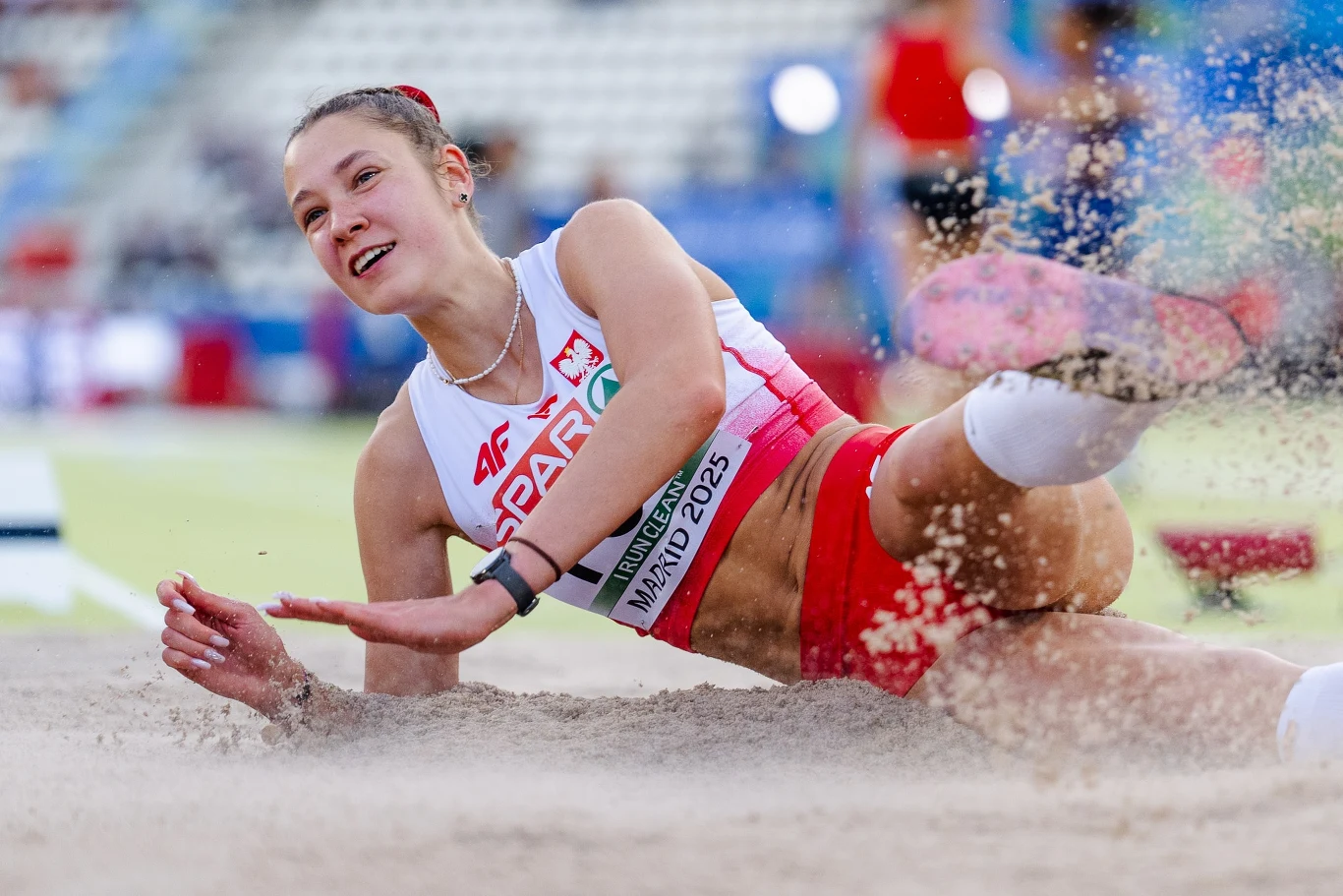 Polska lekkoatletka w trakcie lądowania w piaskownicy podczas konkursu skoku w dal na stadionie lekkoatletycznym, w tle rozmazane trybuny i sportowe wyposażenie.