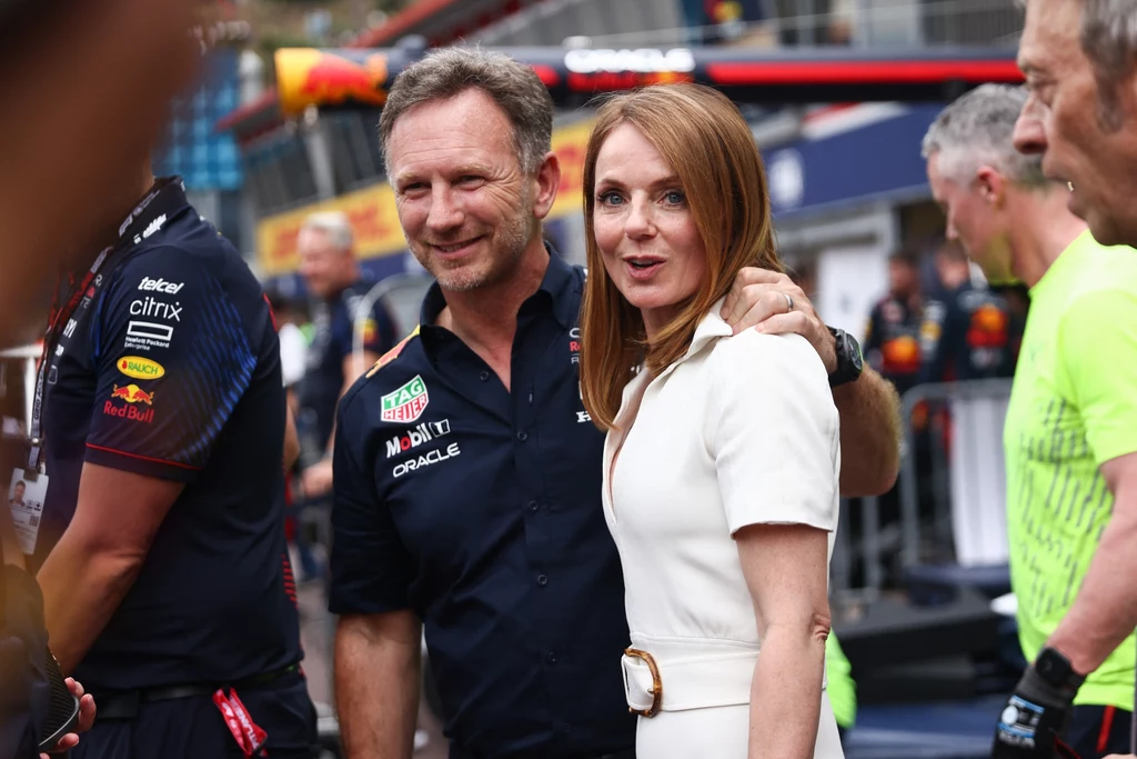 Geri Halliwell i Christian Horner na wyścigu F1 Mężczyzna w koszulce zespołu wyścigowego stoi z uśmiechem obok kobiety w białej sukience, otaczając ją ramieniem. W tle widać zamazane sylwetki innych osób i elementy toru wyścigowego.