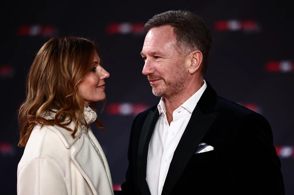 Geri Halliwell i Christian Horner Mężczyzna i kobieta stoją naprzeciwko siebie, patrząc sobie w oczy, ubrane elegancko, kobieta w jasnym płaszczu, mężczyzna w ciemnej marynarce i białej koszuli, ciemne rozmyte tło.