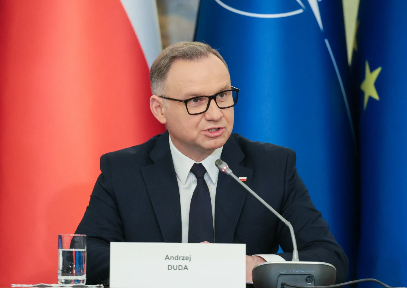 Najnowszy sondaż ocenia prezydenturę Andrzeja Dudy Mężczyzna w garniturze i okularach przemawia na tle flag Polski, Unii Europejskiej oraz NATO, siedząc przy stole z mikrofonem, szklaną wody i tabliczką z nazwiskiem.