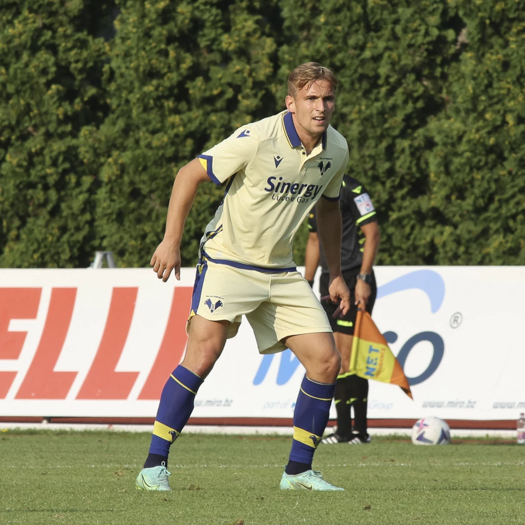 Mateusz Praszelik w barwach Hellasu Verona Piłkarz w jasnym stroju sportowym z logo klubu piłkarskiego oraz sponsora, stoi w lekkim przysiadzie na boisku; w tle widoczny sędzia oraz zielone drzewa, fragmenty banerów reklamowych i piłka do gry.
