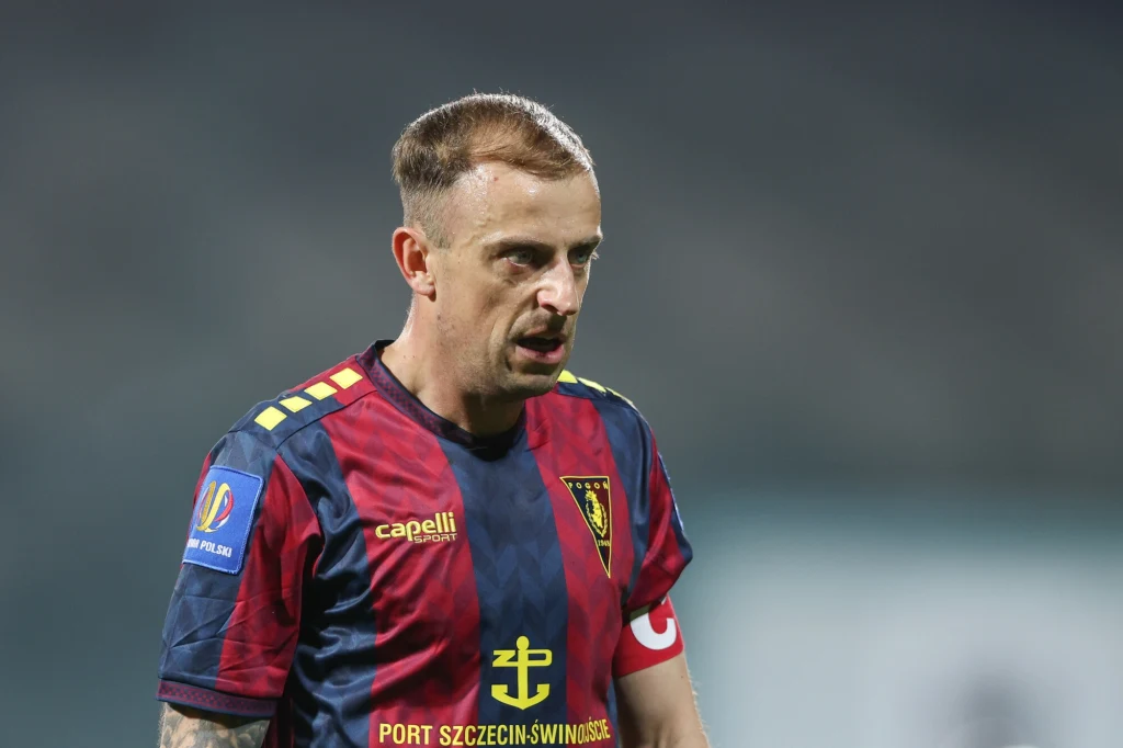 Kamil Grosicki Piłkarz w bordowo-granatowej koszulce z żółtymi detalami i naszywką drużyny Pogoń Szczecin oraz opaską kapitańską na lewym ramieniu. Wyraz twarzy świadczący o skupieniu podczas gry, w tle rozmyte trybuny boiska.