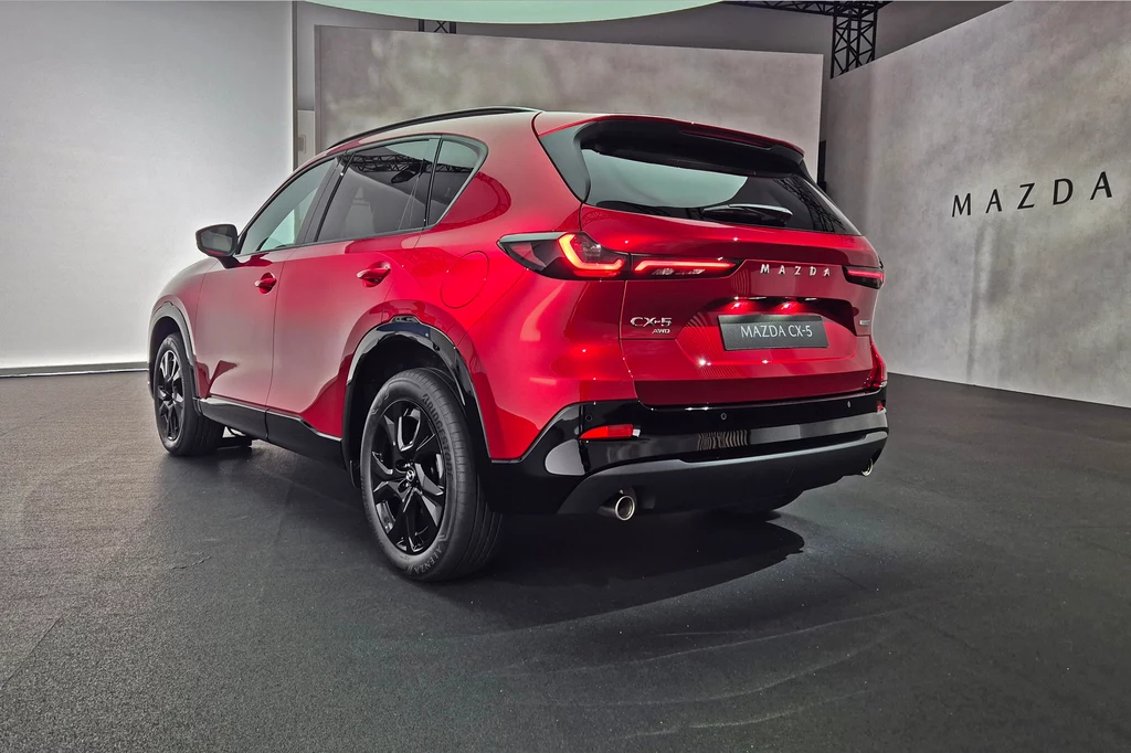 Z tej perspektywy CX-5 przypomina raczej CX-60 niż poprzednika Czerwony samochód Mazda CX-5 stoi w jasnym, nowoczesnym pomieszczeniu ekspozycyjnym, widoczny jest jego tył z wyrazistymi lampami i logo marki.