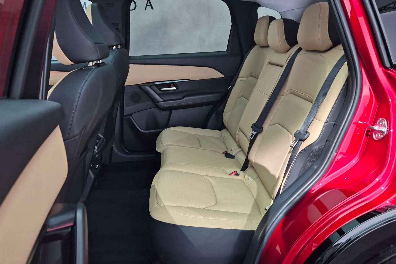 Nowa Mazda CX-5 oferuje sporo miejsca na tylnej kanapie, ale trzeciemu pasażerowi będzie przeszkadzał wysoki tunel środkowy Wnętrze samochodu z widokiem na tylne siedzenia pokryte beżową tapicerką, nowoczesne wykończenie drzwi, pasy bezpieczeństwa oraz część przednich foteli, światło wpada przez boczne okno.