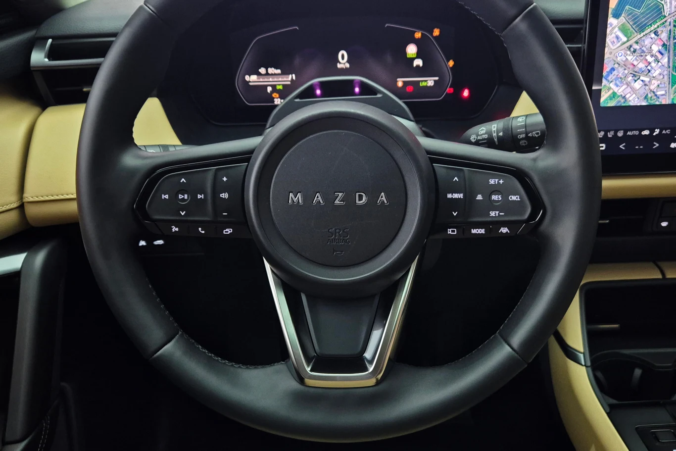 Zamiast znaczka na kierownicy - napis. Nie każdemu się to spodoba. Przyciski wyglądają na dotykowe, ale wciska się je jak klasyczne kierownica samochodu marki Mazda z widocznymi przyciskami sterującymi, cyfrowym panelem wskaźników na desce rozdzielczej oraz fragmentem ekranu nawigacji po prawej stronie, eleganckie wykończenie wnętrza w beżowej tonacji