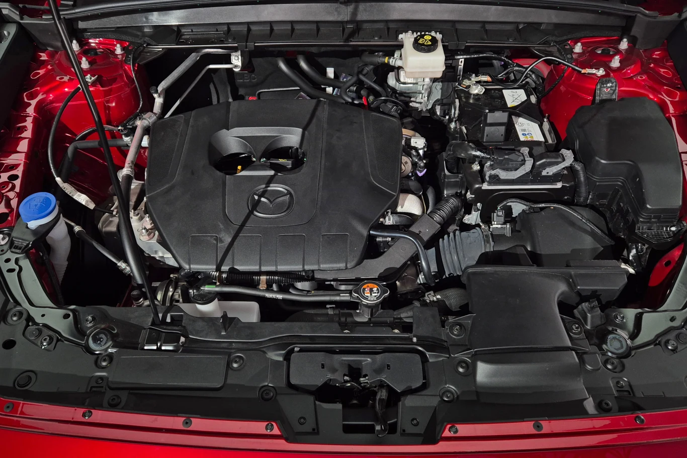 Nowa Mazda CX-5 zachowała 2,5-litrowy, wolnossący silnik, ale rozwija on teraz zaledwie 141 KM Silnik samochodu osadzony w komorze silnikowej, z widocznym logo producenta na pokrywie, otoczony elementami układu chłodzenia, akumulatorem oraz zbiornikiem płynu hamulcowego.