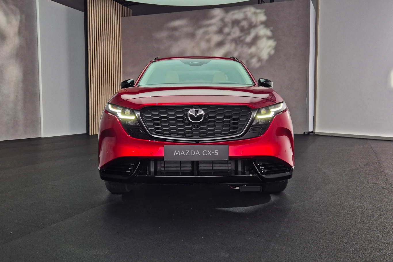 Mazda CX-5 Czerwony samochód osobowy Mazda CX-5 ustawiony przodem, z zapalonymi światłami, na czarnym tle w zamkniętym pomieszczeniu ekspozycyjnym.