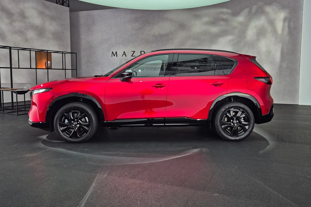 Mazda CX-5 Czerwony samochód typu SUV Mazda ustawiony w nowoczesnym, minimalistycznym wnętrzu z szarymi ścianami i czarnymi regałami po lewej stronie; naprzeciw białej ściany widoczny napis Mazda.