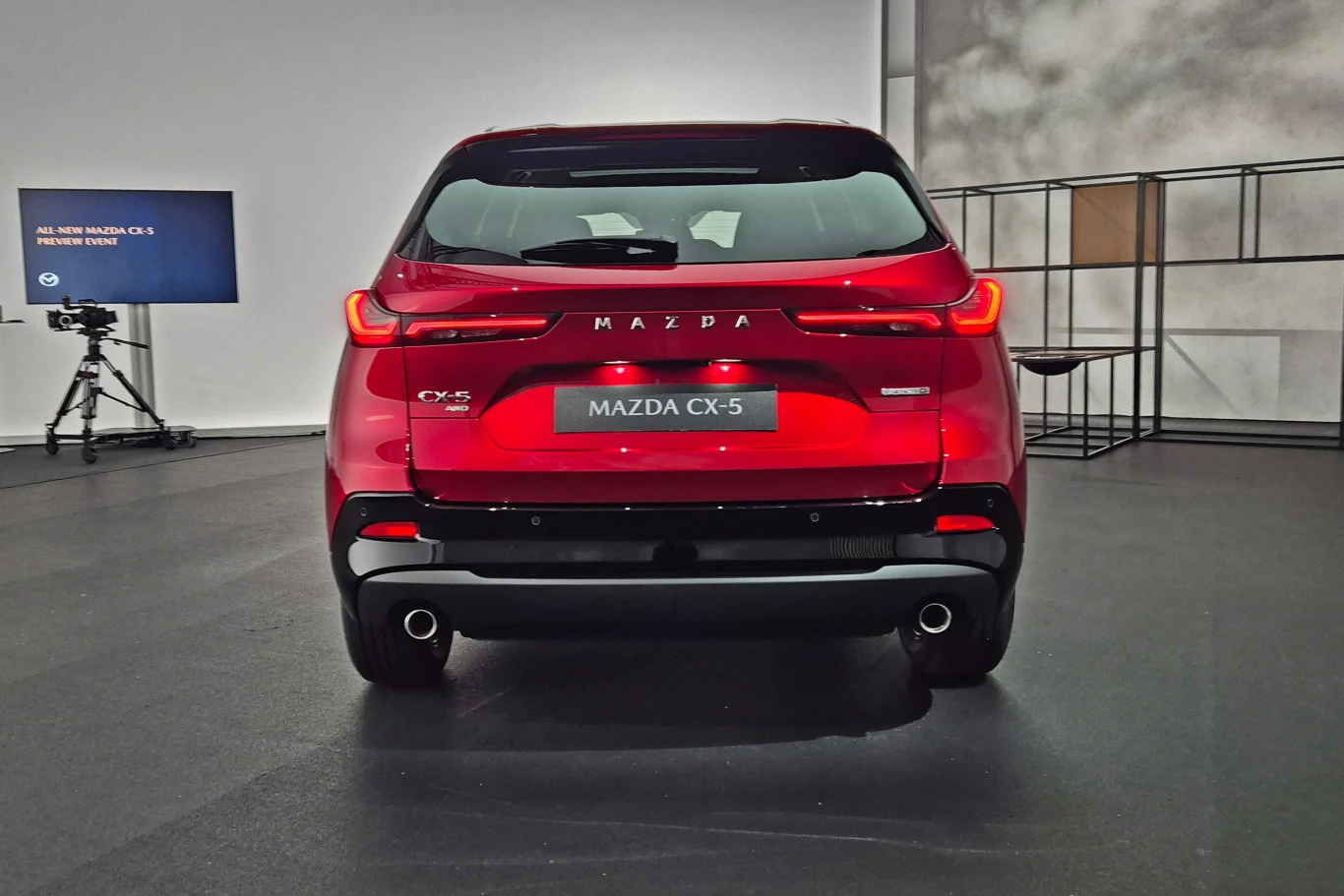 Mazda CX-5 Czerwony samochód Mazda CX-5 widoczny od tyłu w nowoczesnym wnętrzu, obok stoi kamera na statywie i monitor z informacją o premierze modelu.