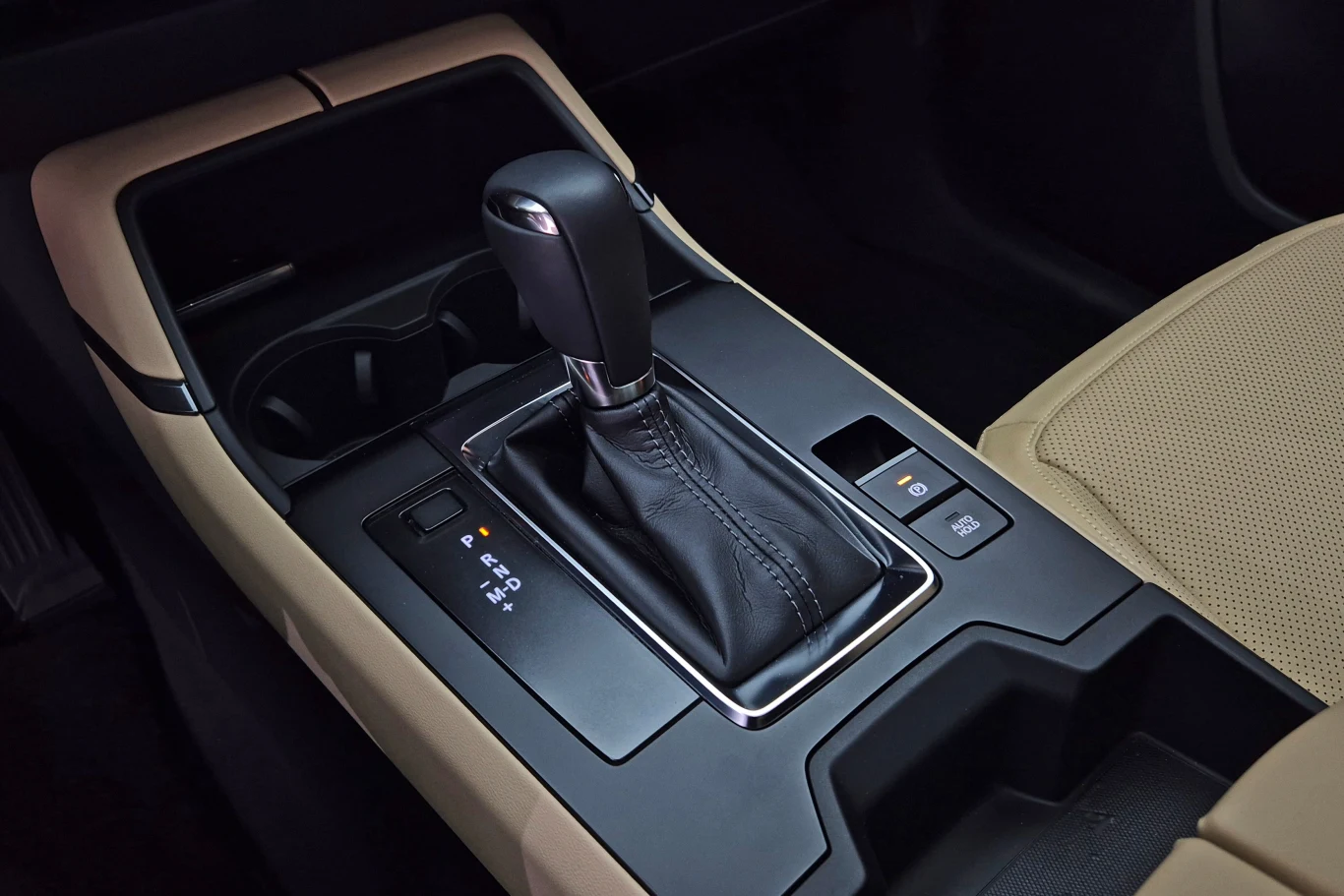 CX-5 występuje tylko z 6-biegowym "automatem". Co ciekawe, choć wnętrze ma nowoczesny i minimalistyczny projekt, pozostawiono klasyczny wybierak skrzyni, zamiast zastosować elektroniczny jak w CX-60. Czarna dźwignia automatycznej skrzyni biegów otoczona beżowo-czarnym wnętrzem samochodu, widoczne przyciski sterujące i uchwyt na kubek w konsoli środkowej.