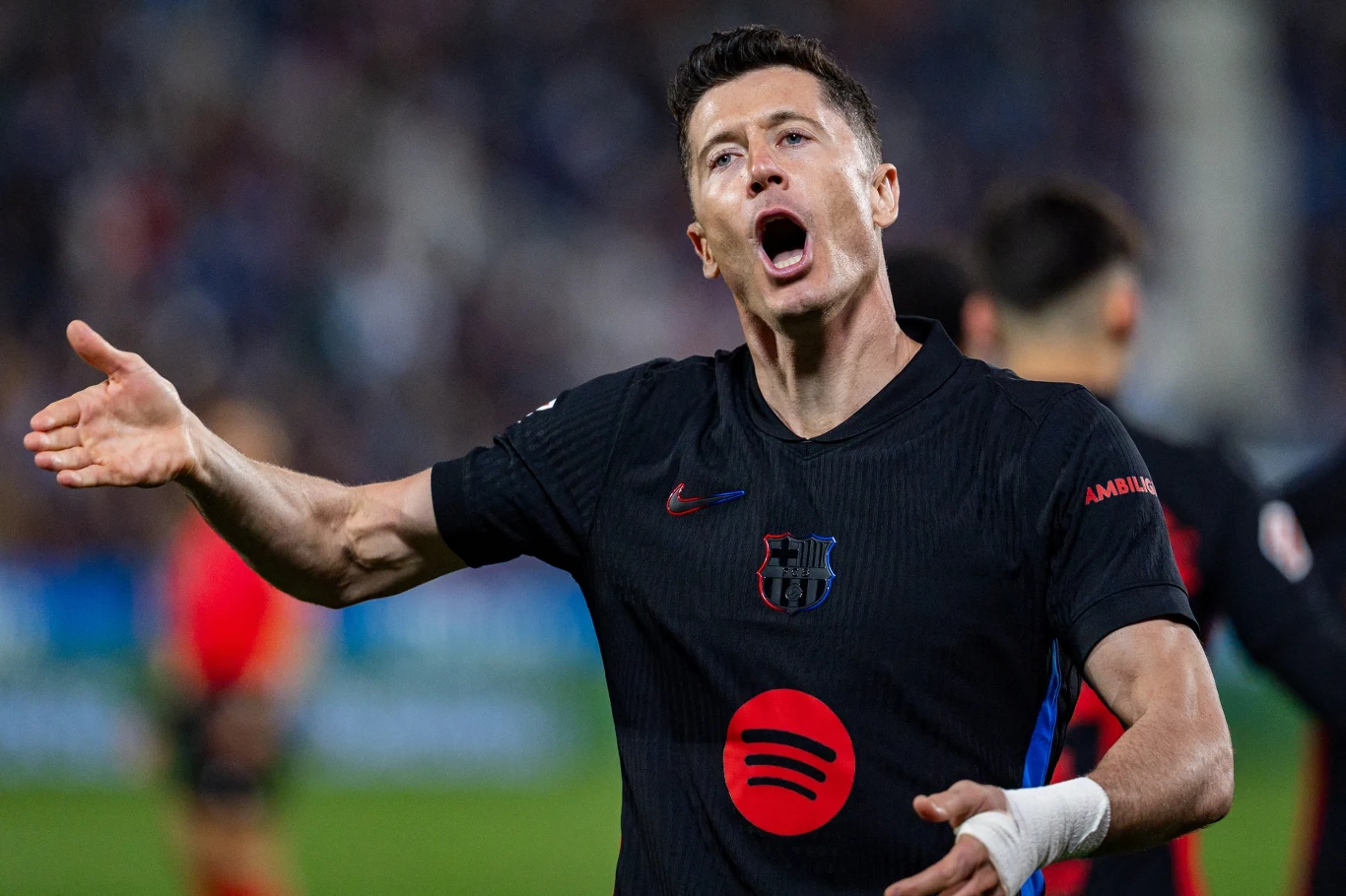 Piłkarz drużyny FC Barcelona w czarnym stroju z charakterystycznym logo, dynamicznie gestykuluje podczas meczu z wyrazem emocji na twarzy; w tle widoczni inni zawodnicy i rozmyta publiczność.