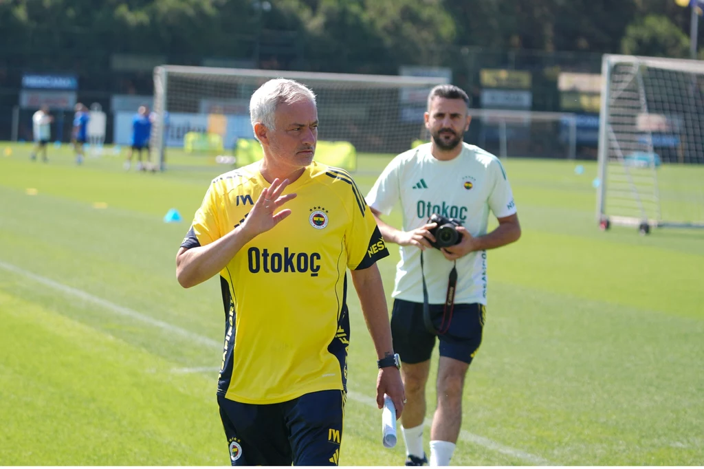 Trener drużyny piłkarskiej w żółtej koszulce przechodzi przez boisko treningowe, w tle widoczny asystent z aparatem fotograficznym oraz bramki i zawodnicy podczas treningu.